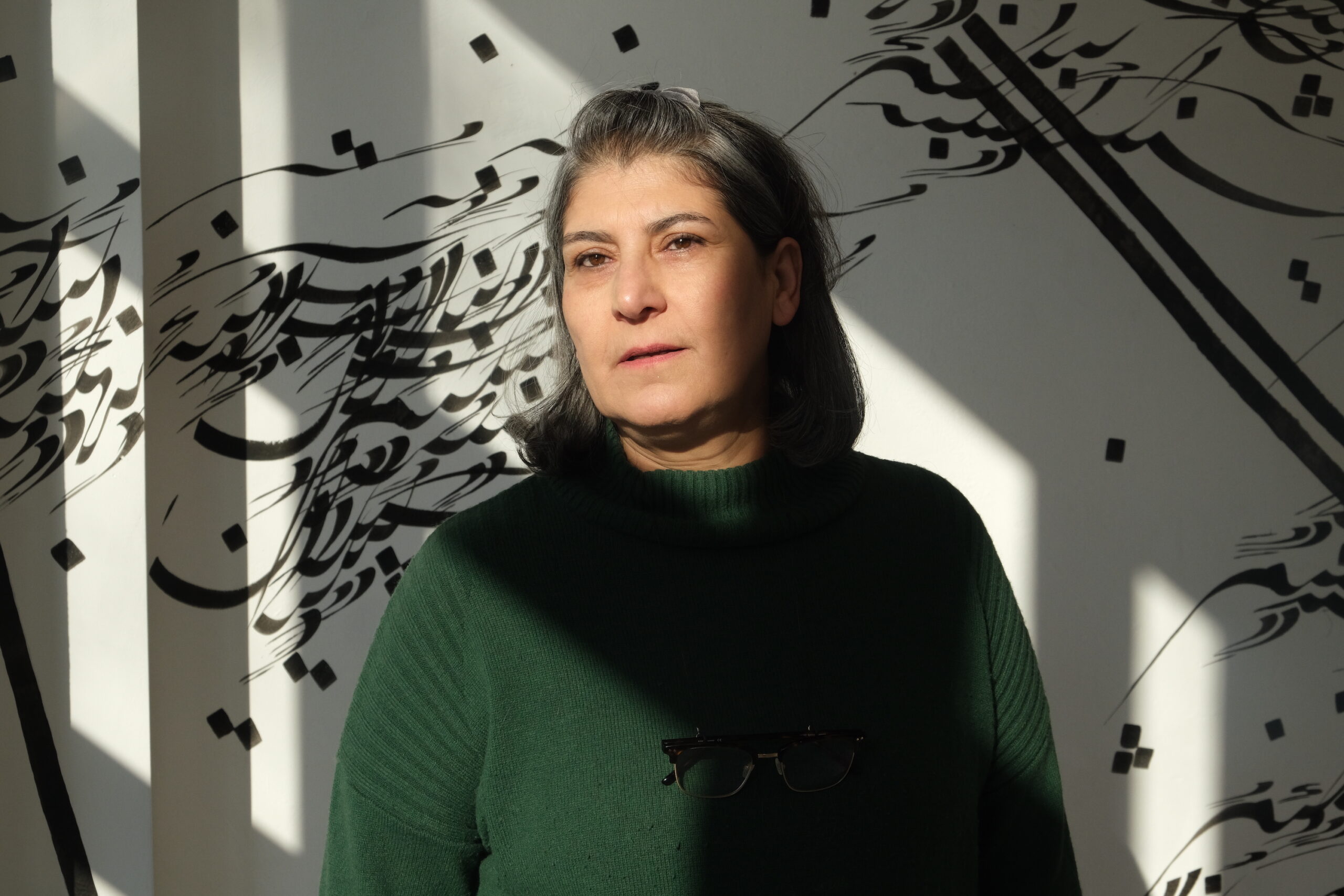 Portrait of Parastou Forouhar in front of her calligraphy installation. She wears a green sweater and stands in direct sunlight.
